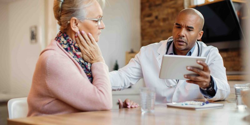 Black doctor reading medical data of mature woman on digital tablet while taking to her during home visit.